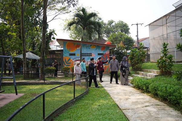 sensei sugimoto kunjungi smart 2 dompet dhuafa Group of people walking along a curved path in a sunny park, with a colorful muralled building on the left and trees in the background.