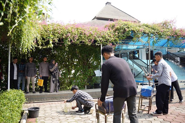 Group of people gathered under a flowering trellis in a shaded courtyard, with a man crouching by a small bucket while others watch.