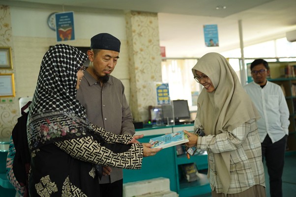 sensei ke smart 6 dompet dhuafa Two women in hijabs exchange a document with a man in a dark cap inside a library, while another man watches in the background.