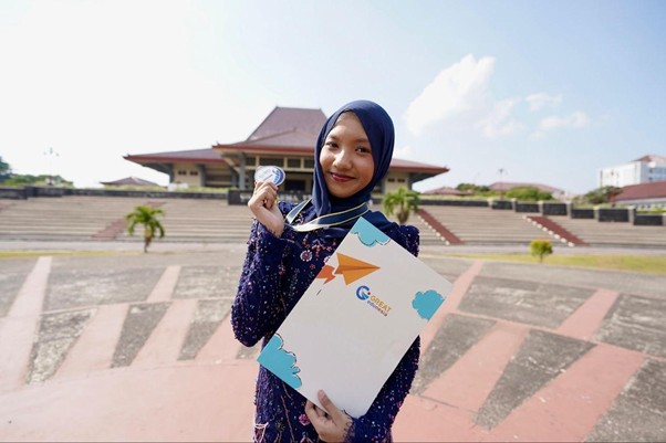 Woman in a blue hijab smiles, holding a certificate and medal outdoors on a campus plaza guarded by stairs and a building in the background.