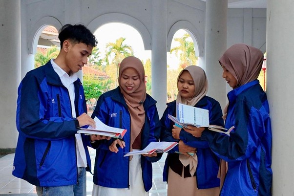 Four students in blue jackets study together under an arched walkway outdoors, two wearing hijabs and one taking notes in a notebook.