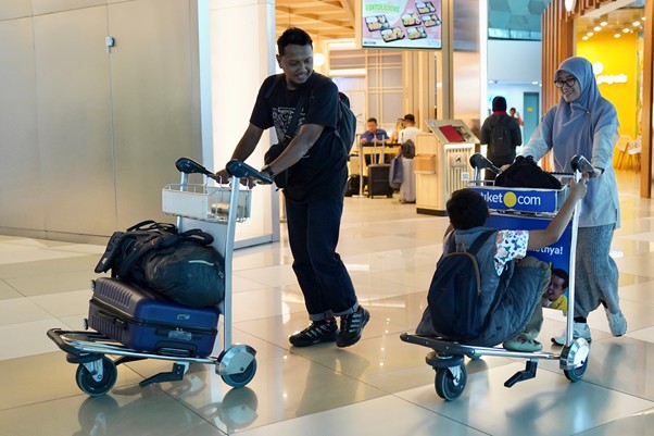 sosokronggodanherman1 dompet dhuafa Man pushes a luggage cart loaded with bags through an airport while a woman wearing a hijab pushes a small two-person luggage scooter carrying a child in front of a shop area.
