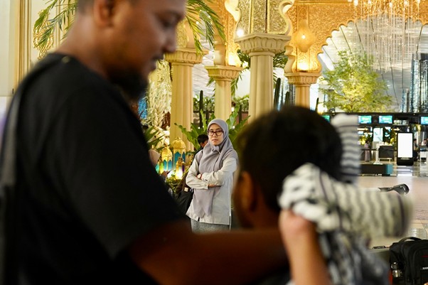 sosokronggodanherman2 dompet dhuafa Barber trimming a customer’s hair in the foreground while a woman in a hijab watches in an ornate, sunlit lobby background.