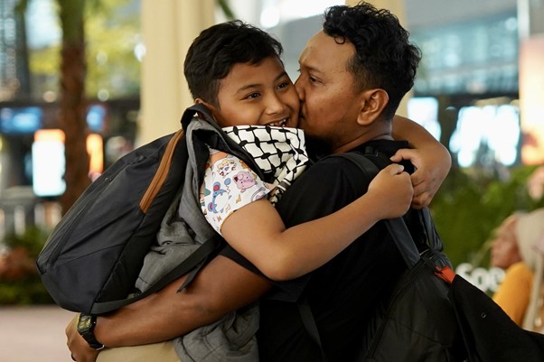 sosokronggodanherman3 dompet dhuafa Parent and child sharing a warm hug in a public indoor space, backpacks on their shoulders.