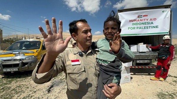 sosokronggodanherman4 dompet dhuafa Aid worker in tan uniform waves with a child against a relief truck banner for Palestine aid effort.