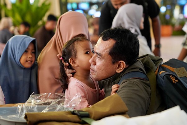 sosokronggodanherman5 dompet dhuafa A man with a backpack crouches as a young girl kisses him on the cheek in a crowded indoor area.