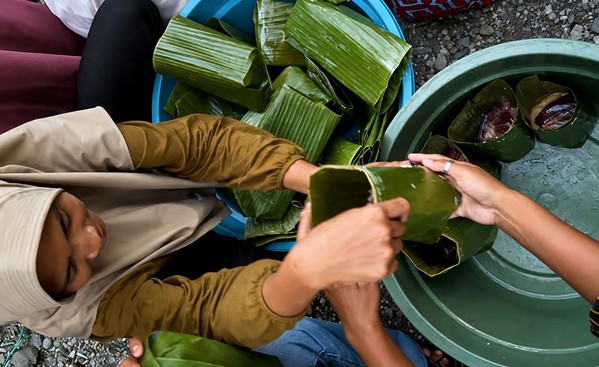 Two people exchange a folded banana leaf parcel while others pile green banana-leaf bundles in a blue tub; a green basin with water nearby.