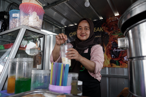 umkm cileungsi 1 dompet dhuafa Woman in a hijab behind a street-food cart, preparing a colorful drink with ice while colorful straws and syrup jars sit nearby.