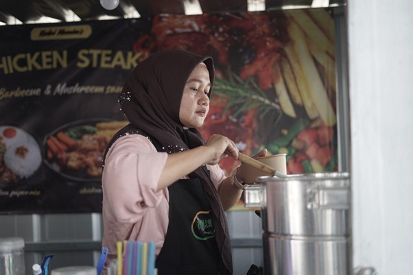 umkm cileungsi 2 dompet dhuafa Woman wearing a hijab and apron cooks at a street food stall, ladling from a pot into a bowl while a large 'Chicken Steak' banner looms in the background.