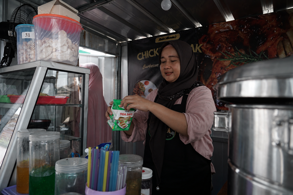 umkm cileungsi 5 dompet dhuafa Female street vendor wearing a hijab pours a drink from a green package at a busy food stall, with colorful straws on the counter in front.