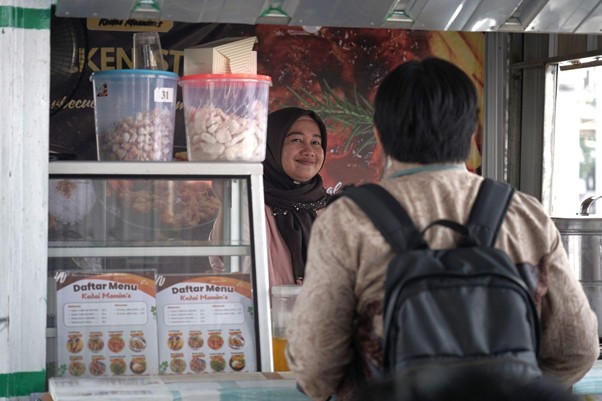 umkm cileungsi 6 dompet dhuafa Female vendor in a hijab smiles at a customer with a backpack at a street food stall, jars of snacks on display and a menu visible behind the glass.