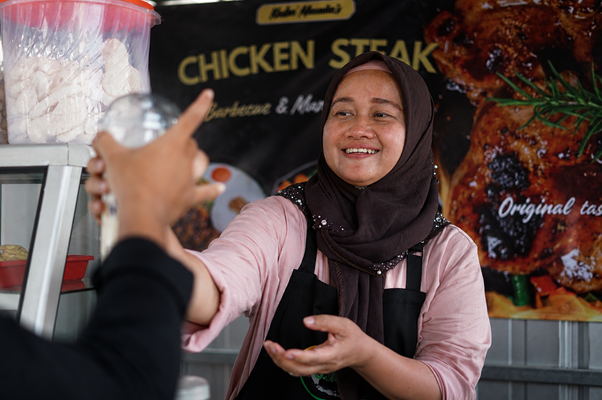 umkm cileungsi 6 dompet dhuafa Female vendor in a hijab hands a wrapped order to a customer at a chicken steak stall.
