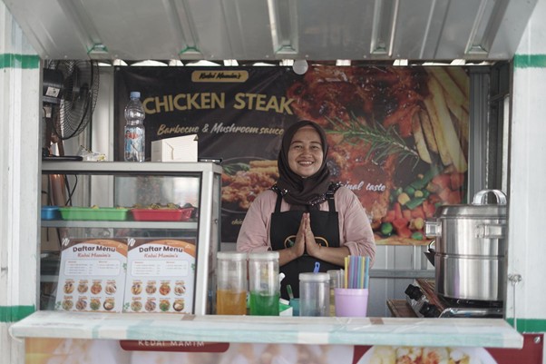 umkm cileungsi 7 dompet dhuafa Smiling woman in a hijab stands behind a street food stall counter with a 'Chicken Steak' banner in the background, hands pressed together in greeting.