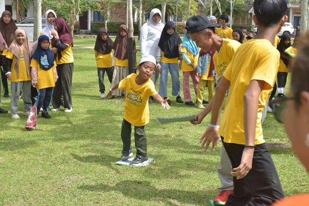 A young boy in a yellow shirt balances on a blue board on a grassy field while a group of children and adults in yellow shirts watch nearby.