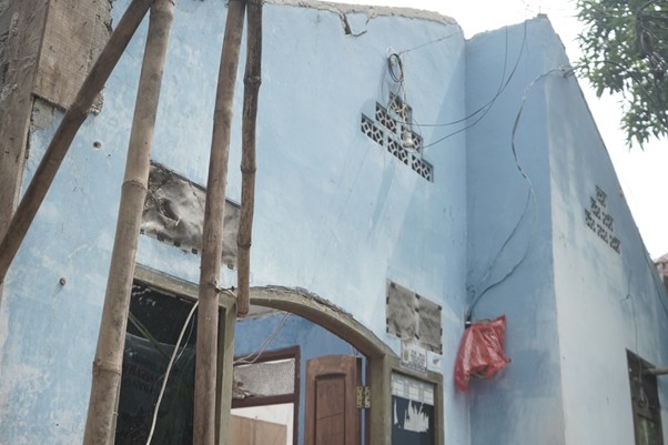 zakatptadev3 dompet dhuafa Blue, partially destroyed building with exposed wires and damaged walls; wooden poles leaning against the facade and an open doorway revealing a cluttered interior.