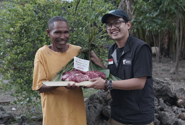 Two men outdoors smile as one hands a banana-leaf platter of raw meat to the other in a donation ceremony.