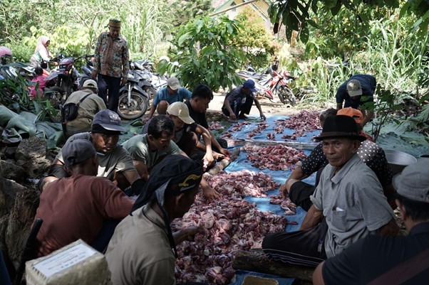 pertumbuhan thk 2026 10 dompet dhuafa Group of people seated around blue tarps covered with heaps of raw meat in a rural outdoor setting, as they sort and process the meat together.
