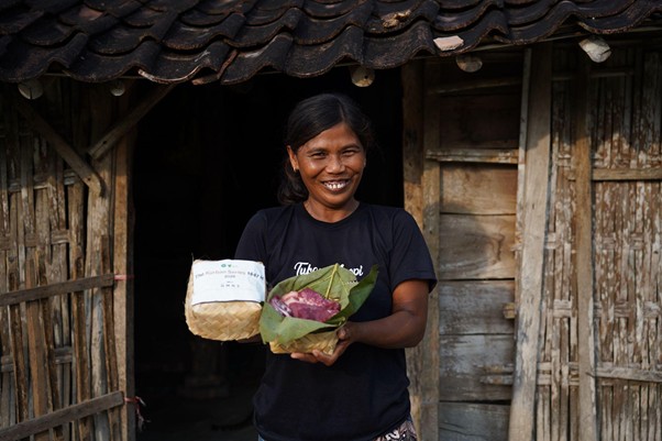 pertumbuhan thk 2026 19 dompet dhuafa Smiling woman standing in a weathered wooden doorway, holding a wrapped package and a bouquet of leaves.