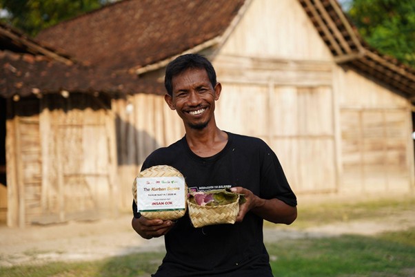 pertumbuhan thk 2026 21 dompet dhuafa Smiling man in a black shirt stands in front of wooden huts, holding a white boxed item and a woven basket.