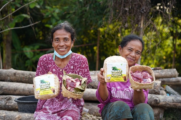pertumbuhan thk 2026 22 dompet dhuafa Two smiling women sit on logs outdoors, each holding baskets with meat and a packaged food item, masks pulled down under their chins.