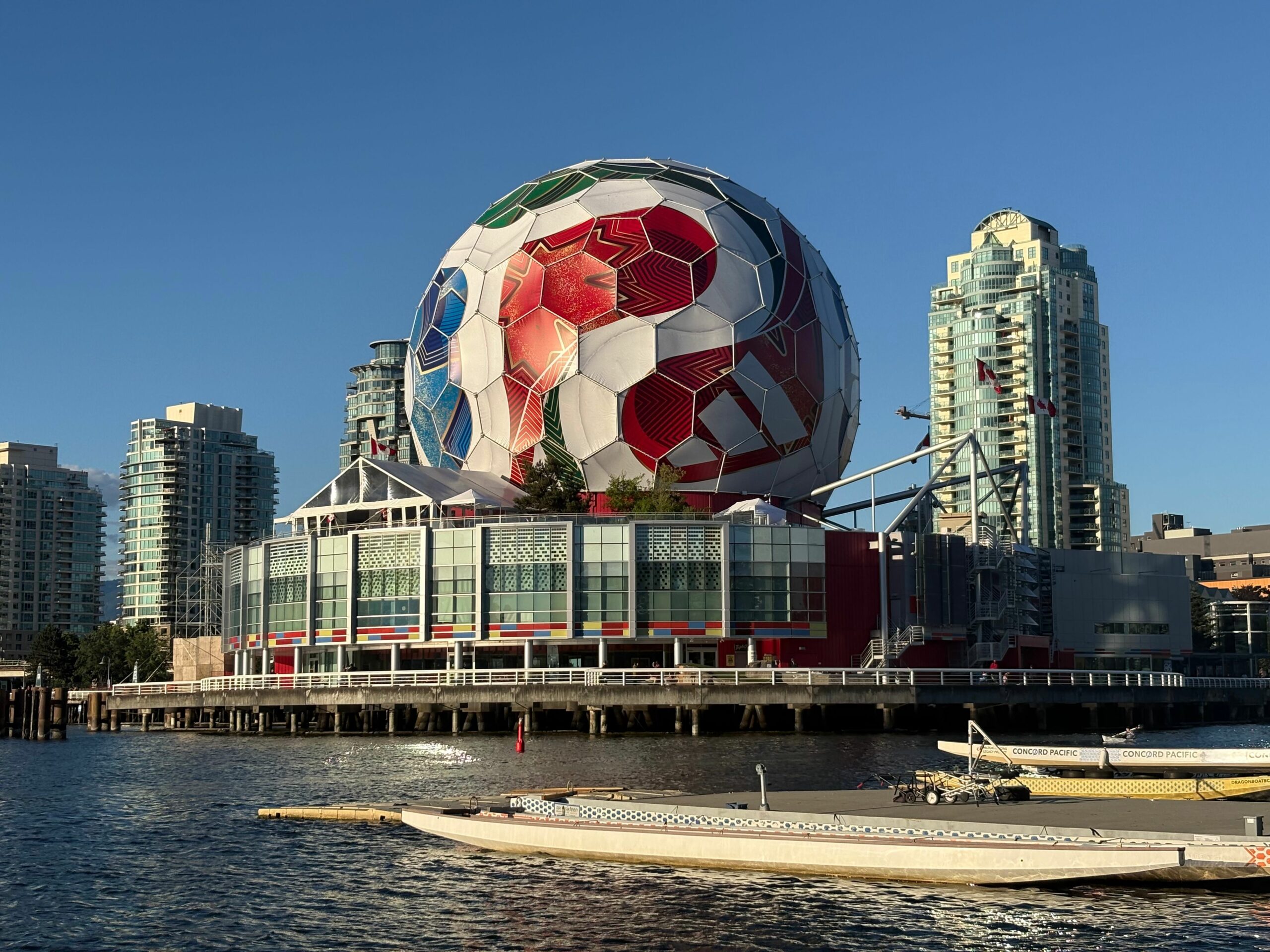 Large colorful geodesic sphere sculpture by the waterfront, with red, green, and white panels, set against modern glass skyscrapers and a clear blue sky.