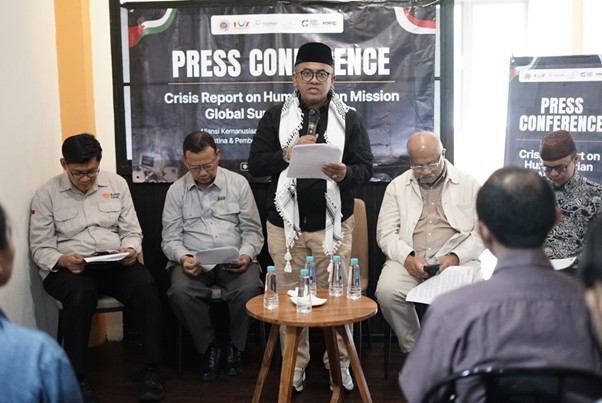 pressconfozgsf204 1 dompet dhuafa Man in a black cap and scarf speaks into a microphone at a press conference, flanked by four panelists seated, with a 'PRESS CONFERENCE' banner in the background.