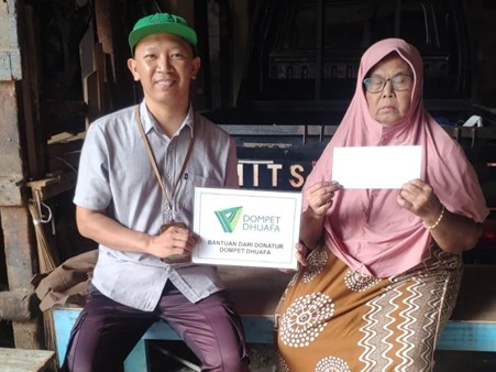 Man in a green cap hands a Dompet Dhuafa donation box to an elderly woman in a pink hijab, who holds a white envelope.