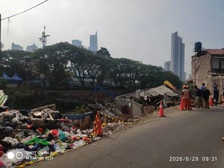 bantuansosialdarilpm2 dompet dhuafa Trash and debris piled along a riverside road while people stand nearby; city skyline in the background.