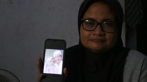 Woman in a hijab and glasses holds up a smartphone showing a photo of two smiling people in white clothing on the screen.