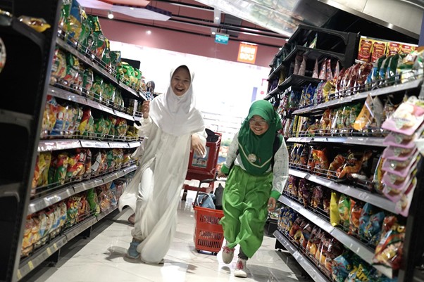 belanja bareng yatim trans studio mall cibubur 1 dompet dhuafa Two smiling girls in hijabs run down a supermarket aisle between snack shelves, one in white and one in green, with a red shopping basket nearby.