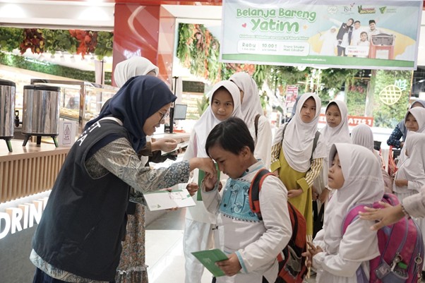 belanjabarengyatimtransstudiomallcibubur12 dompet dhuafa A woman in a hijab hands a small item to a boy as several girls in white uniforms and hijabs observe in a busy food court or market area.