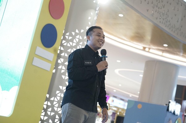 belanjabarengyatimtransstudiomallcibubur7 dompet dhuafa Man in a dark shirt holding a microphone, speaking on a stage at an indoor event with decorative panels nearby
