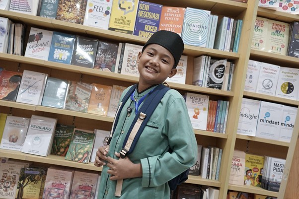 besteam gramedia 1 dompet dhuafa Smiling boy in a green traditional outfit with a backpack in a bookstore, shelves filled with books behind him.