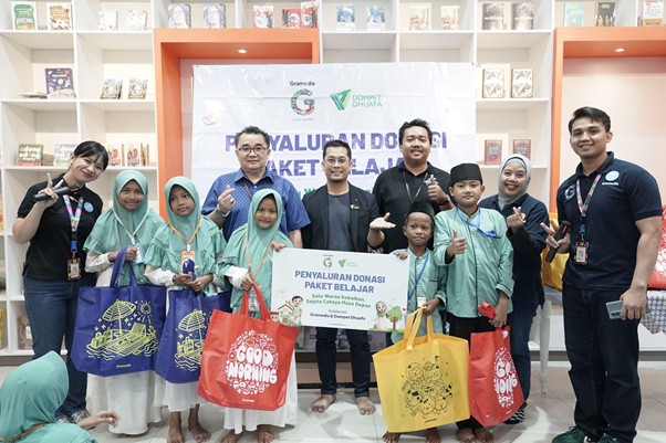 Group of children in green headscarves and adults posing with donation bags at a learning-packs donation event in a library setting.