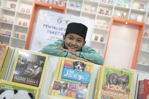 Smiling boy in a green shirt and black cap rests his chin on a colorful display of children's books in a bright bookstore.