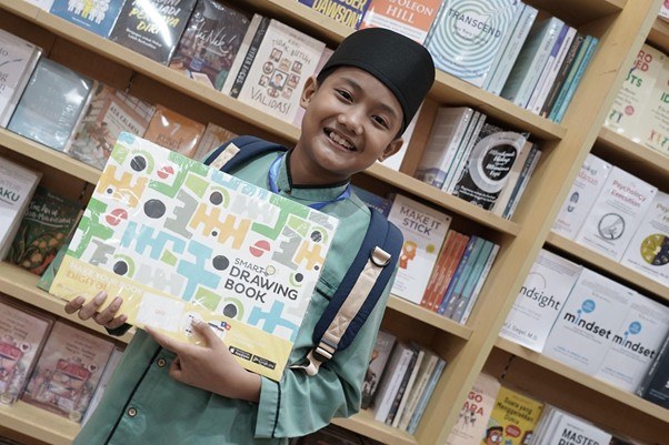 besteam gramedia 3 dompet dhuafa Smiling young girl in a hijab and backpack holding a large colorful'Smart Drawing Book' in a bookstore.