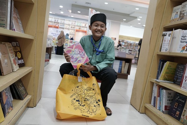 Smiling boy in a green outfit kneels in a bookstore, holding a pink illustrated book and a yellow cartoon‑shaped tote bag on his lap.