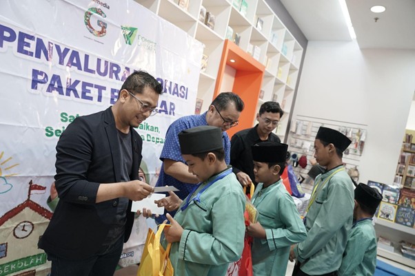 Adult in a blazer hands a gift to a boy in a green uniform during a ceremony, with other children waiting and a banner in Indonesian in the background.
