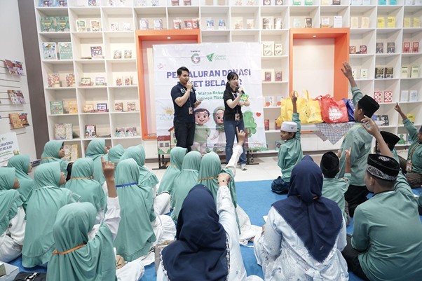 besteam gramedia 7 dompet dhuafa Audience seated on the floor in teal hijabs watches two presenters on a stage with a banner behind them; some raise hands in the crowd at a community event.