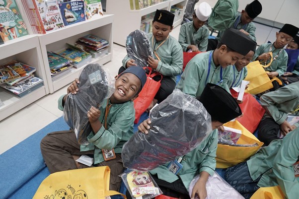 Group of smiling Muslim schoolchildren in green uniforms sit on the library floor with large gift bags and backpacks, surrounded by books.