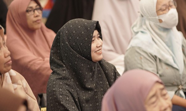 Smiling woman in a dark patterned hijab seated among a diverse audience of Muslim women.