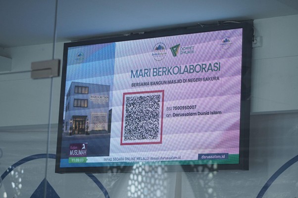 Digital display promoting collaboration to build a mosque, featuring the slogan ‘MARI BERKOLABORASI’ and a QR code with logos and a building image.