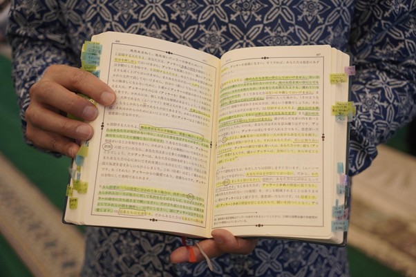 Open Japanese book with extensive yellow highlights and many colored sticky tabs, held by a person in a blue patterned outfit.
