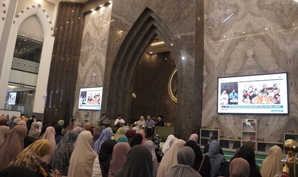 Crowded auditorium with women wearing hijabs watching a panel discussion on stage in a marble-walled hall.