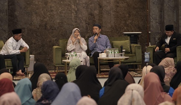 Panel discussion on stage with four speakers in front of an audience of women wearing headscarves; two speakers hold microphones and converse on stage.