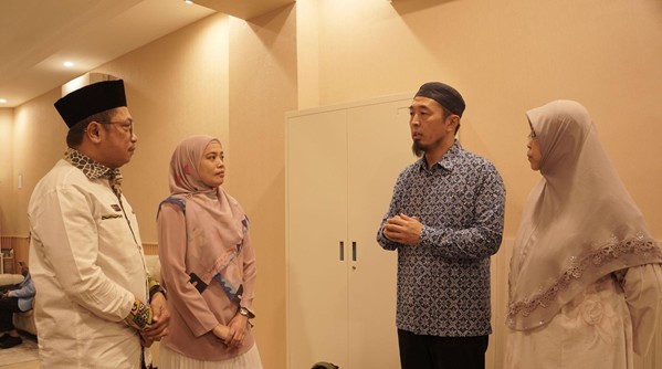 Four adults in a hallway conversation; two men wearing traditional caps and long-sleeve shirts, two women in hijabs listening to the discussion.