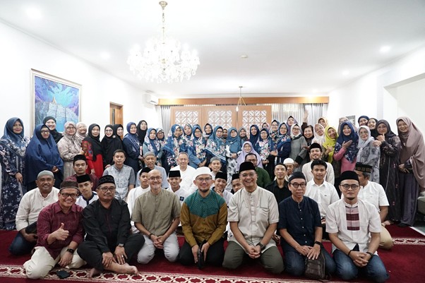 Large group photo of men and women in a bright room with a chandelier, carpet, and windows; many women wearing hijabs, all posing together for the shot.