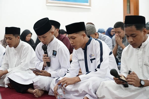 Group of men in white robes and black caps sit on a prayer carpet; one holds a microphone while another checks his phone, with others praying in the background.