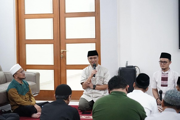 A man in a black cap speaks into a microphone while seated on a stage, surrounded by attendees listening in a prayer circle in a bright room with wooden doors behind him.
