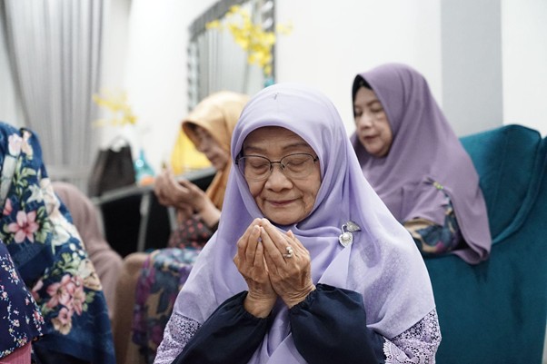 Elderly woman in a lavender hijab prays with cupped hands during a gathering, others seated in the background.